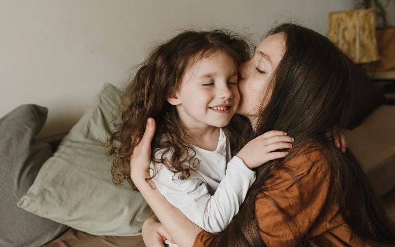 Mother gently kisses smiling daughter during warm embrace.