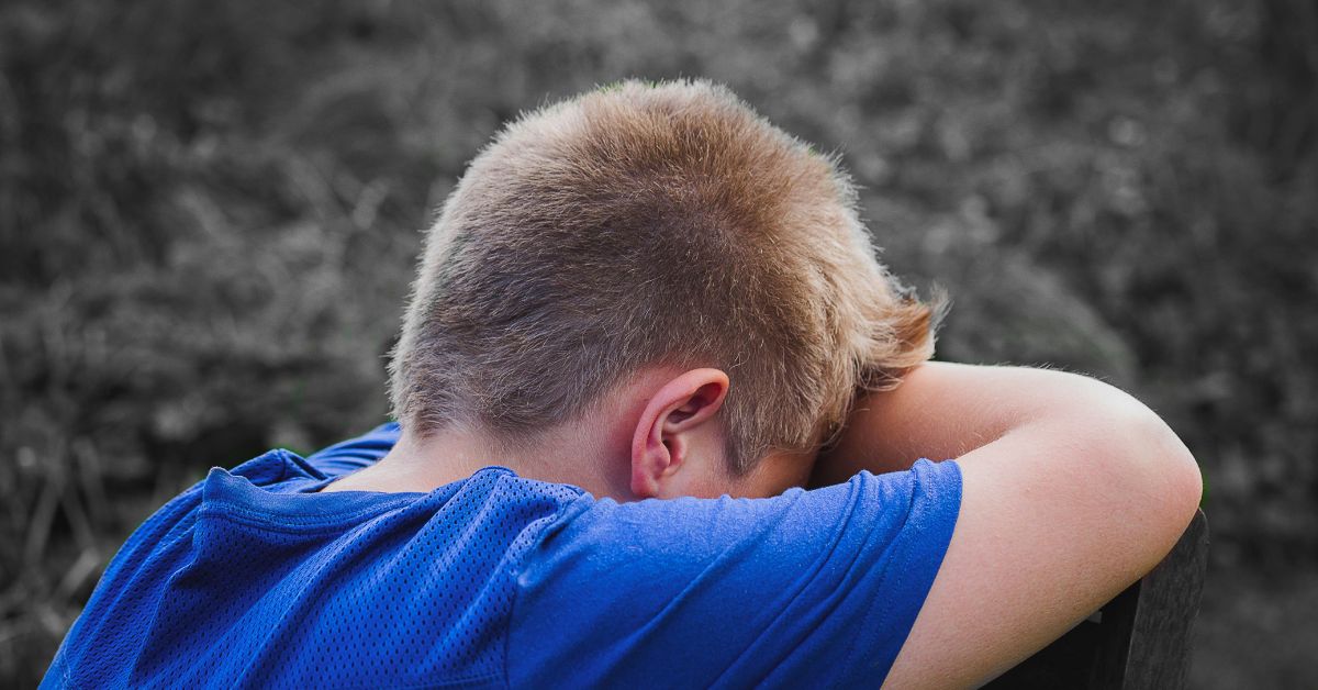 A young boy in a blue shirt rests his head on his arms outdoors, appearing sad and contemplative